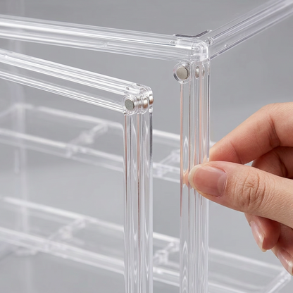 A close-up of a hand assembling a clear acrylic organizer box, showing the transparent plastic frame and corner connectors against a light gray background.