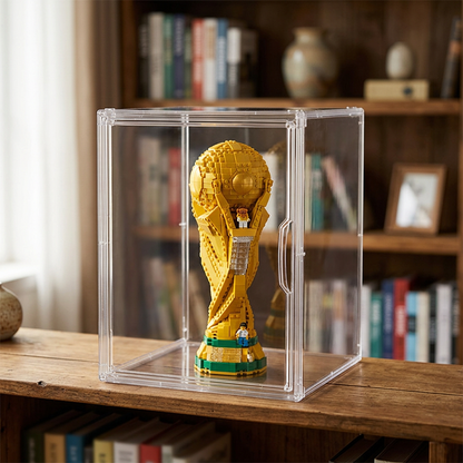 A clear acrylic display case containing a golden soccer trophy figure, placed on a wooden shelf in front of a blurred bookshelf background.