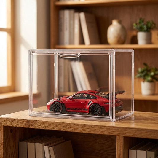A red sports car model displayed inside a clear acrylic storage box on a wooden shelf, with a softly blurred indoor background featuring books and plants.